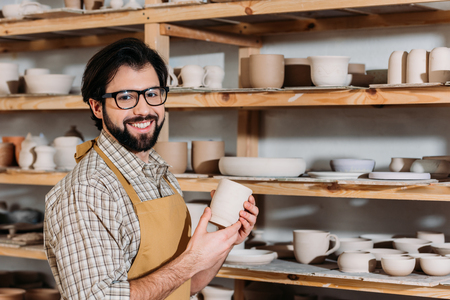 Smiling potter holding ceramic cup in workshop with dishware on shelvesの写真素材