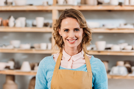 Beautiful cheerful woman in apron standing in pottery workshopの写真素材