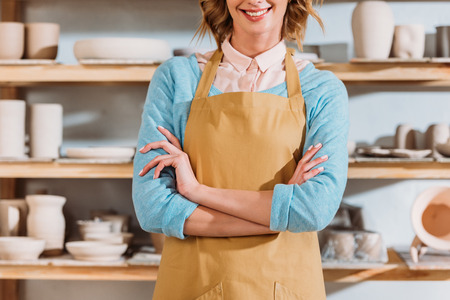 Cropped view of potter with crossed arms standing near shelves with ceramic dishwareの写真素材