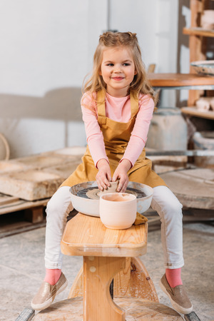 Blonde smiling kid making ceramics on pottery wheel in workshopの写真素材