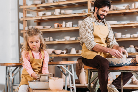 Kid making ceramic pot on pottery wheel with teacher near in workshopの写真素材
