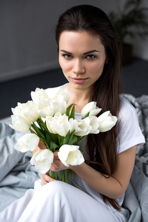 Pretty young woman holding bouquet of tulips in bedの写真素材