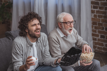 adult son and senior father cheering on baseball team and watching game at home