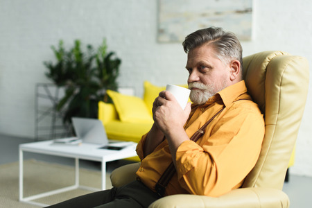 smiling bearded senior man drinking coffee from cup while sitting on armchair at home