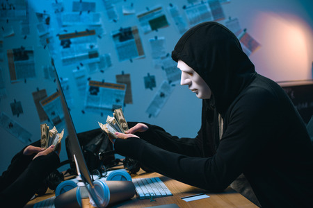 side view of hooded hacker in mask counting stolen money at his workplace