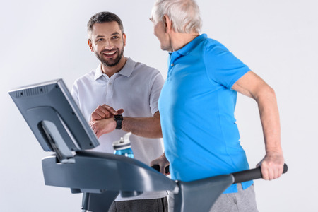 smiling rehabilitation therapist assisting senior man exercising on treadmill isolated on greyの写真素材