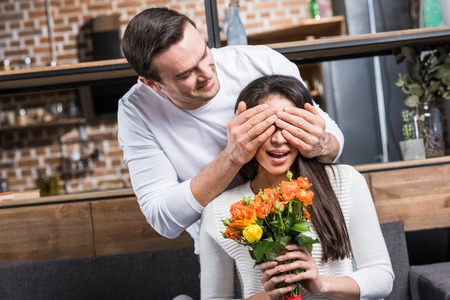 happy man closing eyes to girlfriend holding bouquet of flowers at homeの写真素材