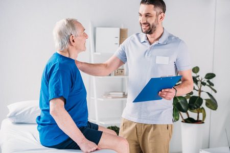 smiling rehabilitation therapist with notepad and senior man on massage tableの写真素材