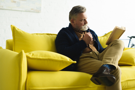 bearded senior man sitting on yellow couch and reading bookの写真素材