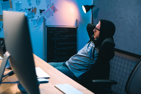 young hooded hacker relaxing on chair in front of his workplace