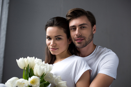 Smiling man and woman with flowers tenderly embracing in bedroomの写真素材