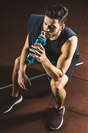 High angle view of sportsman drinking water from bottle and sitting on barbell in gymの写真素材