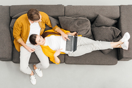 top view of woman lying on knees of boyfriend and working with laptop on cozy couch at homeの写真素材