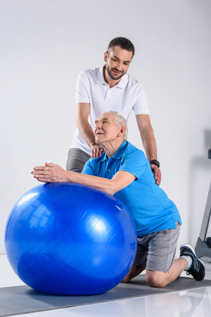 rehabilitation therapist making massage to senior man on fitness ballの写真素材