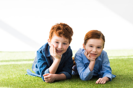 happy redhead siblings lying together on grass and smiling at camera on grey