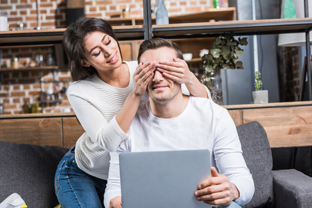 smiling african american woman closing eyes to caucasian boyfriend using laptop at homeの写真素材