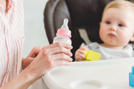 Cropped view of mother holding feeding bottle and infant daughter sitting in baby chair with plastic blocksの写真素材