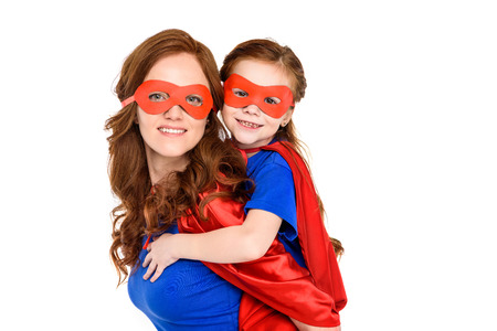 super mother piggybacking adorable daughter in mask and cloak and smiling at camera isolated on...