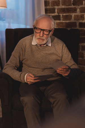 senior  man sitting in armchair, holding old photos and looking awayの写真素材