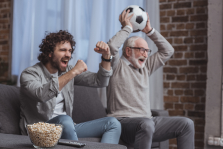 adult son and senior father watching football game and showing yes signs at homeの写真素材