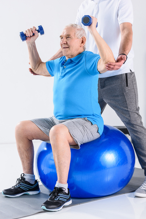 cropped shot of rehabilitation therapist assisting senior man exercising with dumbbells on fitness ball isolated on greyの写真素材