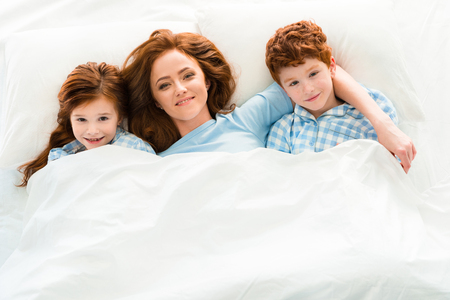 top view of happy mother with cute redhead kids lying in bed and smiling at cameraの写真素材