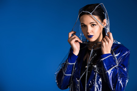 Young girl with blue lipstick wearing transparent raincoat isolated on blue backgroundの写真素材