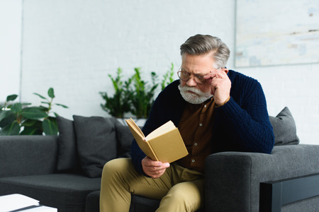 stylish senior man in eyeglasses sitting on sofa and reading book at homeの写真素材