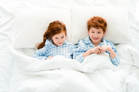 beautiful happy redhead kids in pajamas lying together in bed and smiling at cameraの写真素材