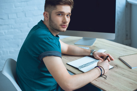 handsome young freelancer taking notes and looking at camera while working at homeの写真素材