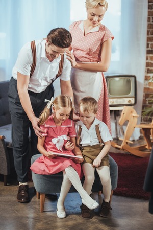 happy parents looking at cute little kids using digital tablet, 1950s style