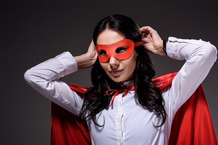 attractive super businesswoman in red cape tying mask isolated on blackの写真素材