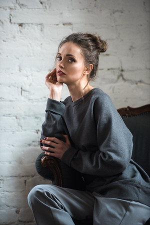 portrait of beautiful brunette woman in grey trendy clothes sitting on armchair and looking at cameraの写真素材