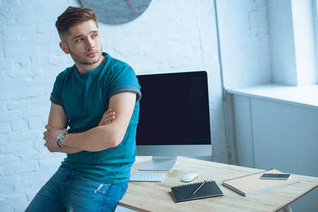 handsome young man with crossed arms sitting on table and looking away while working at homeの写真素材