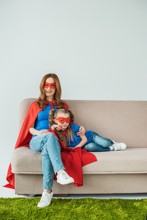 super mother and daughter in masks and cloaks sitting on sofa and smiling at cameraの写真素材