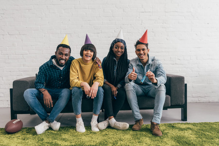 smiling group of young multiethnic friends in party hats sitting on couchの写真素材
