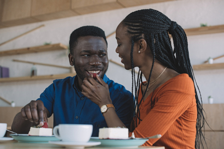 african american woman feeding boyfriend with strawberry at table in cafeの写真素材