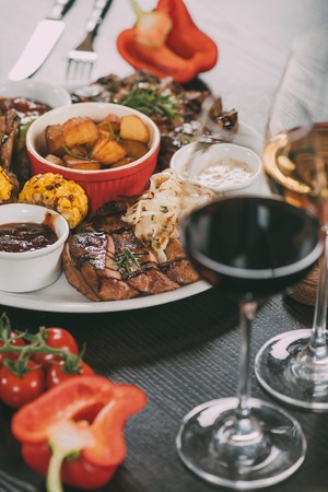 plate with sauces, roasted potatoes, grilled meat with vegetables and glasses of wine on tableの写真素材