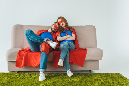 tired couple in superhero costumes sleeping while sitting on sofa on greyの写真素材