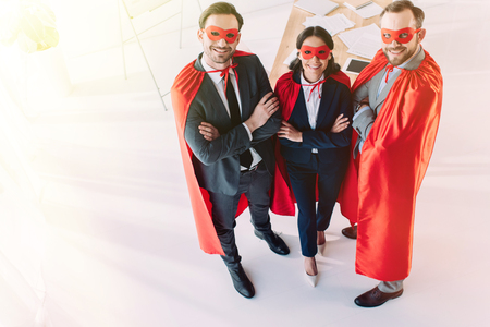 high angle view of super businesspeople in masks and capes with crossed arms looking at camera in officeの写真素材