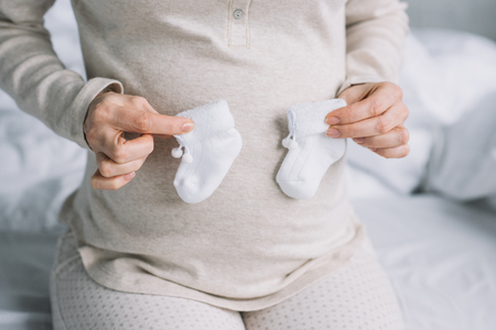 cropped image of pregnant woman touching belly with newborn shoes in bedroomの写真素材
