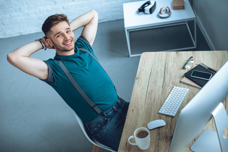 high angle view of handsome young freelancer sitting with hands behind head and smiling at cameraの写真素材