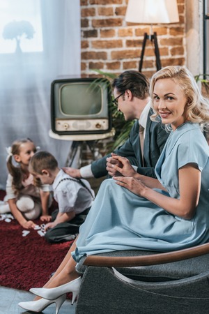 happy 50s style woman smiling at camera while sitting on sofa with husband and kids playing with...