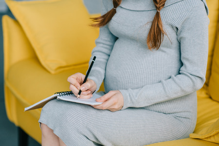 cropped image of pregnant woman writing something to notebook on sofaの写真素材