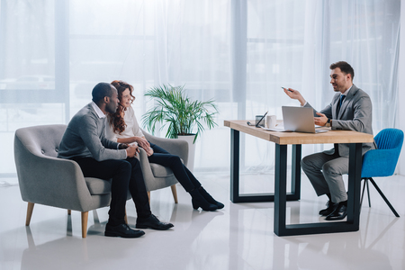 Interracial couple sitting in armchairs and estate agent at table with laptopの写真素材