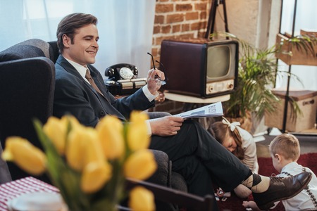 smiling man in suit reading newspaper in armchair while kids playing with domino tiles behind, 1950s...