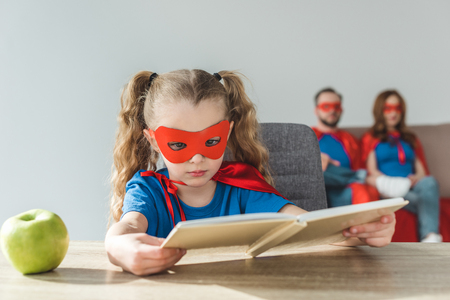 girl in superhero costume reading book while super parents sitting behindの写真素材