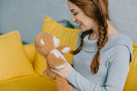 side view of beautiful pregnant woman looking at teddy bear in living roomの写真素材