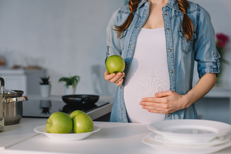 cropped image of pregnant woman holding apple and touching belly at kitchenの写真素材