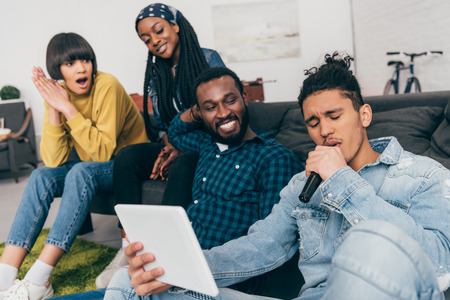 young mixed race man with microphone and digital tablet singing karaoke and his friend sitting besideの写真素材